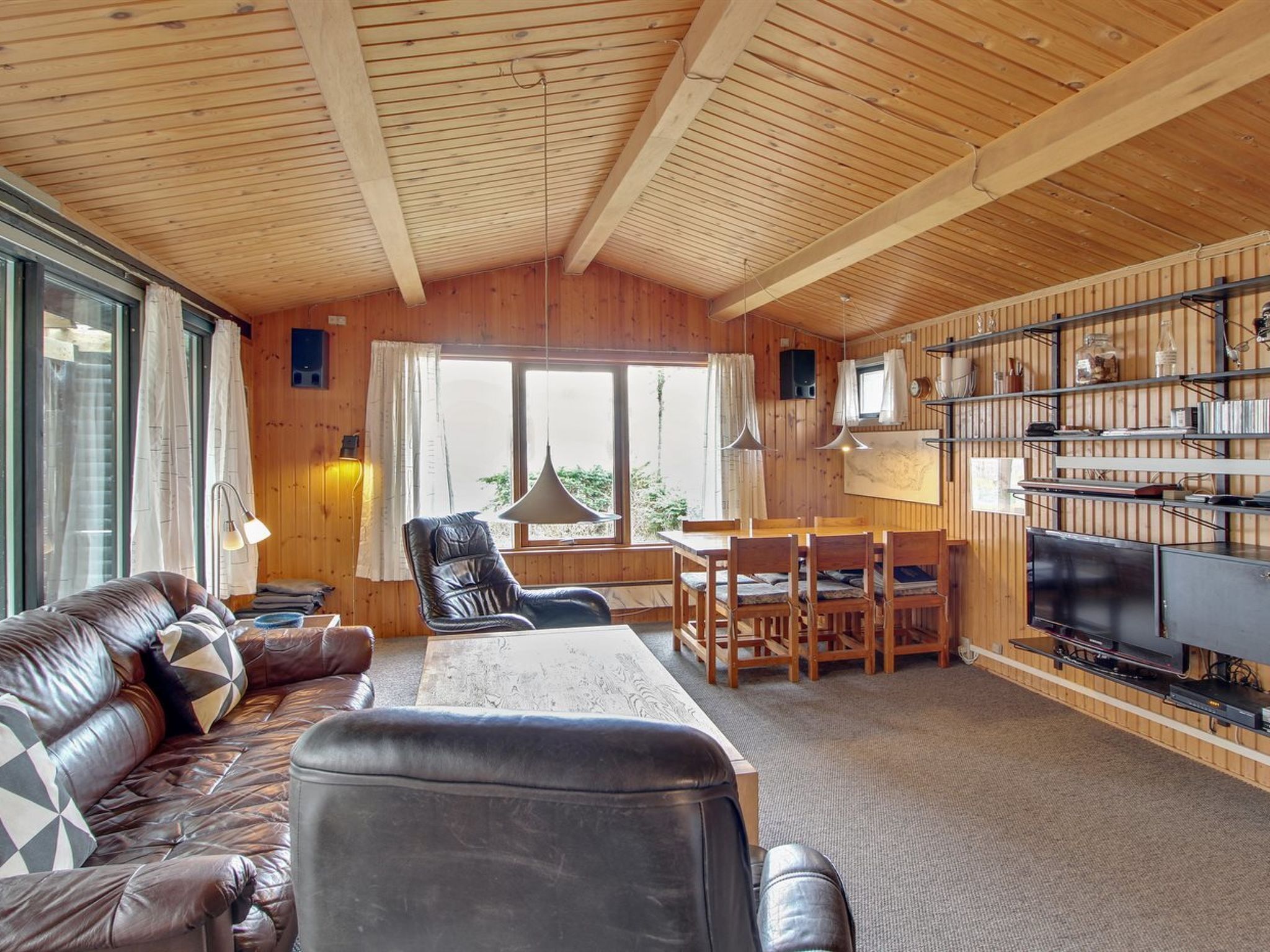 A cozy cabin interior with a wooden ceiling, a leather sofa, a dining table, chairs, a TV, and a bookshelf.