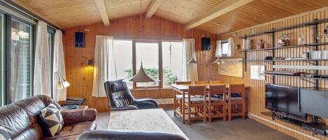 A cozy cabin interior with a wooden ceiling, a leather sofa, a dining table, chairs, a TV, and a bookshelf.