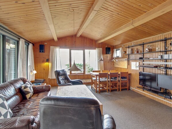 A cozy cabin interior with a wooden ceiling, a leather sofa, a dining table, chairs, a TV, and a bookshelf.