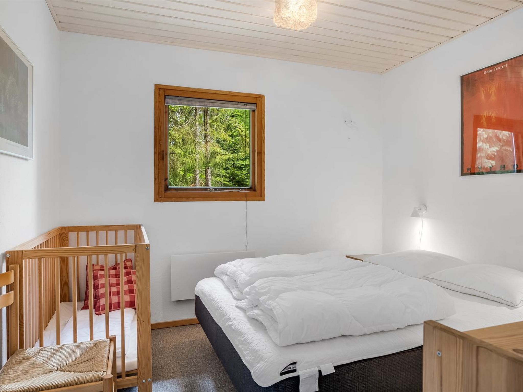 Ein Schlafzimmer mit einem Bett, einem Kinderbett, einem Fenster mit Blick auf einen Wald und einem an der Wand befestigten Kunstwerk.