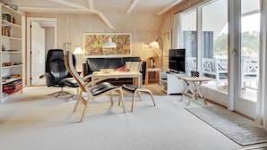 A modern living room with a black leather sofa, wooden chairs, a coffee table, and a TV. There is a bookshelf, a painting, and a large window with a view.
