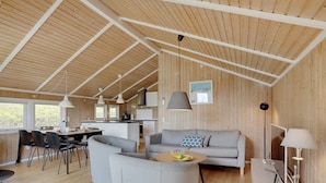 Un'ampia zona giorno con soffitto in legno, un divano grigio, un tavolino da caffè con della frutta e un'area pranzo con sedie e tavolo.