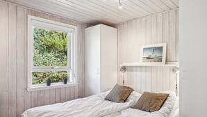 Un dormitorio con una cama, dos almohadas, una ventana con plantas y un cuadro en la pared.