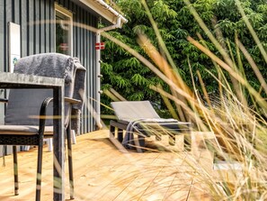 Une terrasse en bois avec un fauteuil de relaxation et une table, un bâtiment avec un revêtement vertical, et un feuillage verdoyant et dense en arrière-plan.