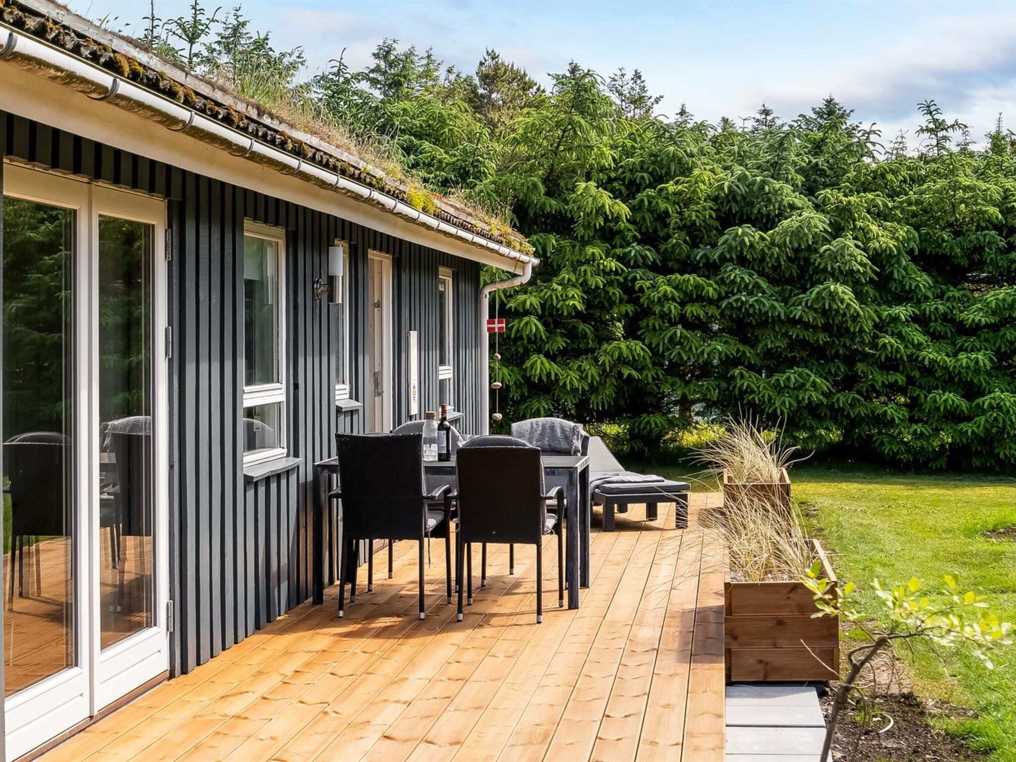 Ett modernt hus med en veranda, utomhusmöbler och en frodig, grön trädgård.