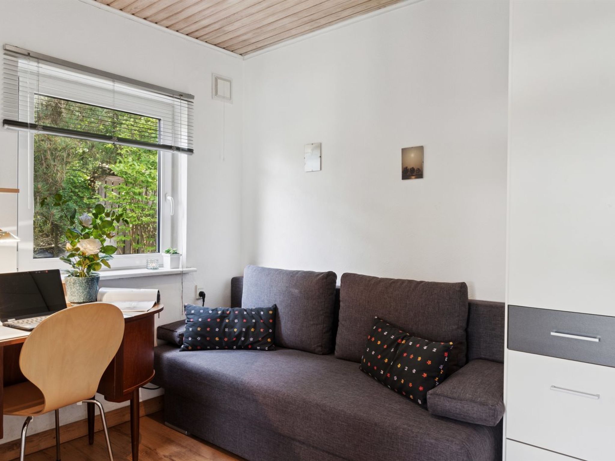 A modern room with a grey sofa, a wooden desk, and a laptop. There is a window with blinds, a plant on the windowsill, and a small wall-mounted light.