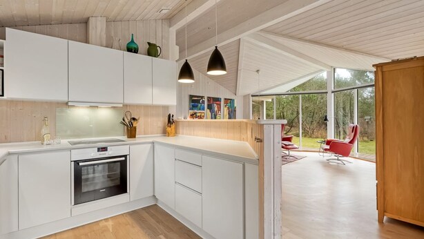 Een moderne keuken met witte kasten, een inbouwoven en hanglampen. Er is een eethoek met een rode stoel en uitzicht op de omgeving.