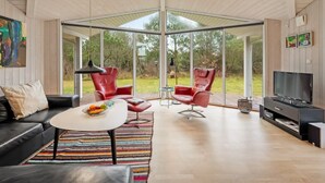 Een ruime woonkamer met een grote glazen deur die uitkomt op een tuin, een zwarte leren bank, rode stoelen, een witte salontafel en een televisie op een zwarte standaard.