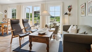 En rummelig opholdsstue med et træbord, to lænestole, en sofa og et spisebord med stole. Store vinduer giver udsigt til det fri.
