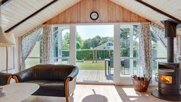 Ett mysigt rum med ett träpanelat tak, en svart lädersoffa, ett runt bord, en öppen spis och utsikt över en trädgård genom stora glasdörrar.