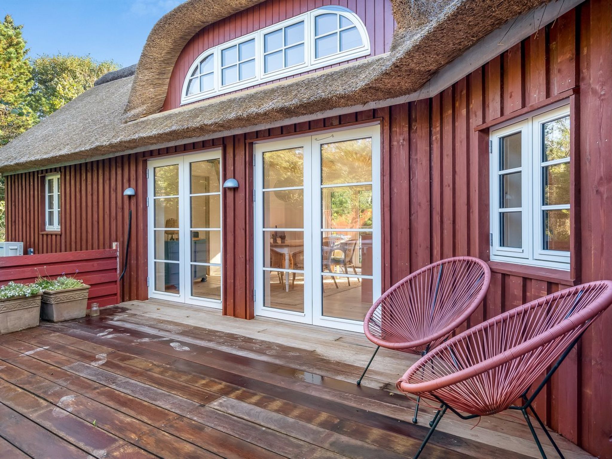 Una casa de madera con techo de paja, grandes puertas correderas de vidrio y una terraza de madera con dos sillas de mimbre.