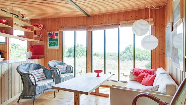 Interno di una baita in legno con zona pranzo, due poltrone in vimini, un divano e ampie finestre.