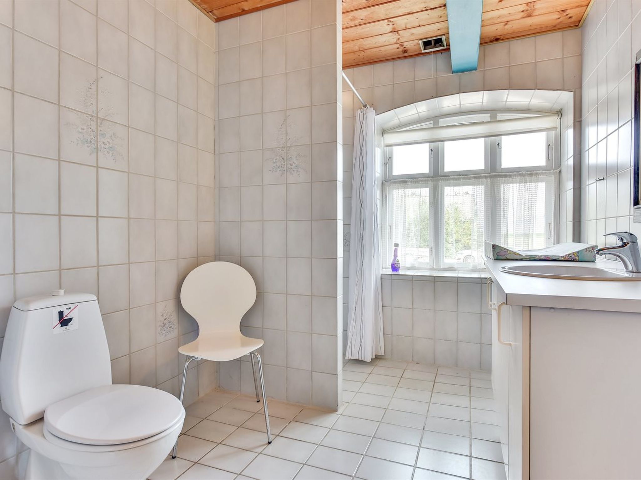 A bathroom with a toilet, a chair, a window, and a sink.
