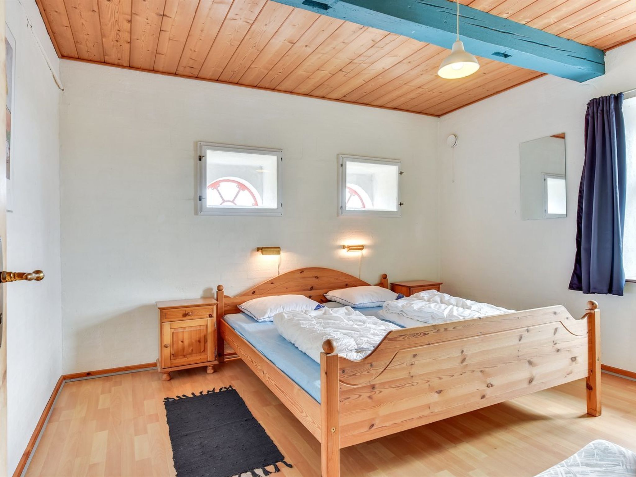 A wooden bed with a headboard, two bedside tables, a black rug, and two windows with curtains.