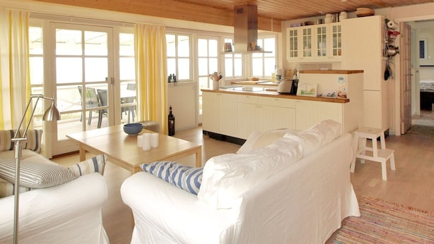 Een ruime woonkamer met houten plafond, een witte bank, een eettafel, en een keuken met witte kasten en een houten aanrechtblad.