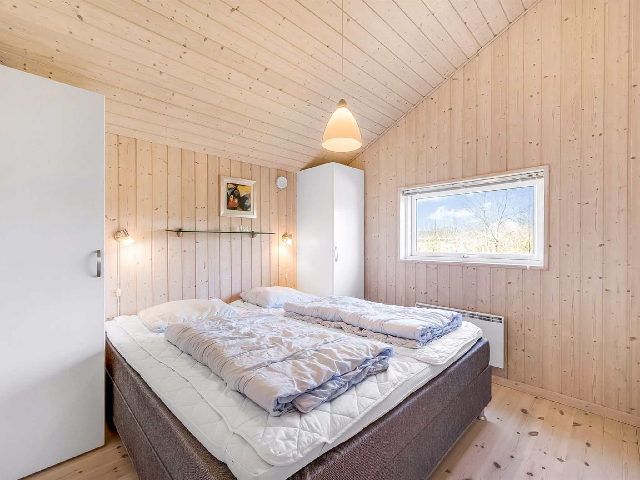Une chambre avec un plafond en bois, un lit avec une literie blanche, une lampe de chevet, une fenêtre donnant sur l’extérieur et une étagère murale avec un tableau.