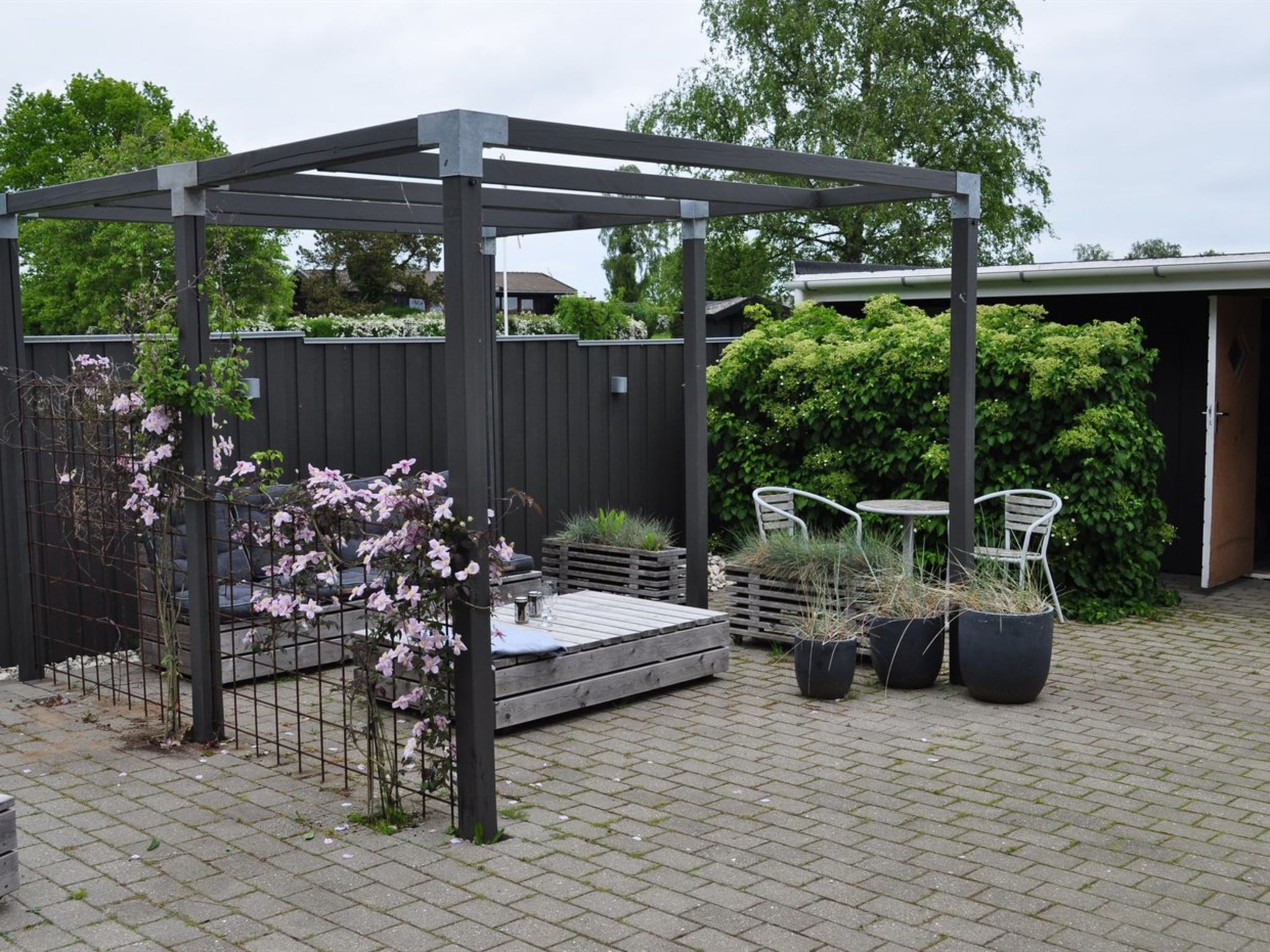 A covered patio area with a wooden pergola, outdoor seating, potted plants, and a brick floor.
