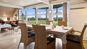 Un comedor con techo alto y ventanas amplias, sillas de mimbre y una mesa lista para comer.