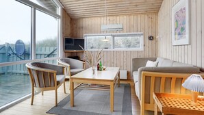 A cozy cabin interior with a wooden ceiling, a dining area with a table and chairs, a sofa, a TV, and a large window offering a view of the outdoors. - "Kalervo" - 100m from the sea by Interhome (Pandrup)
