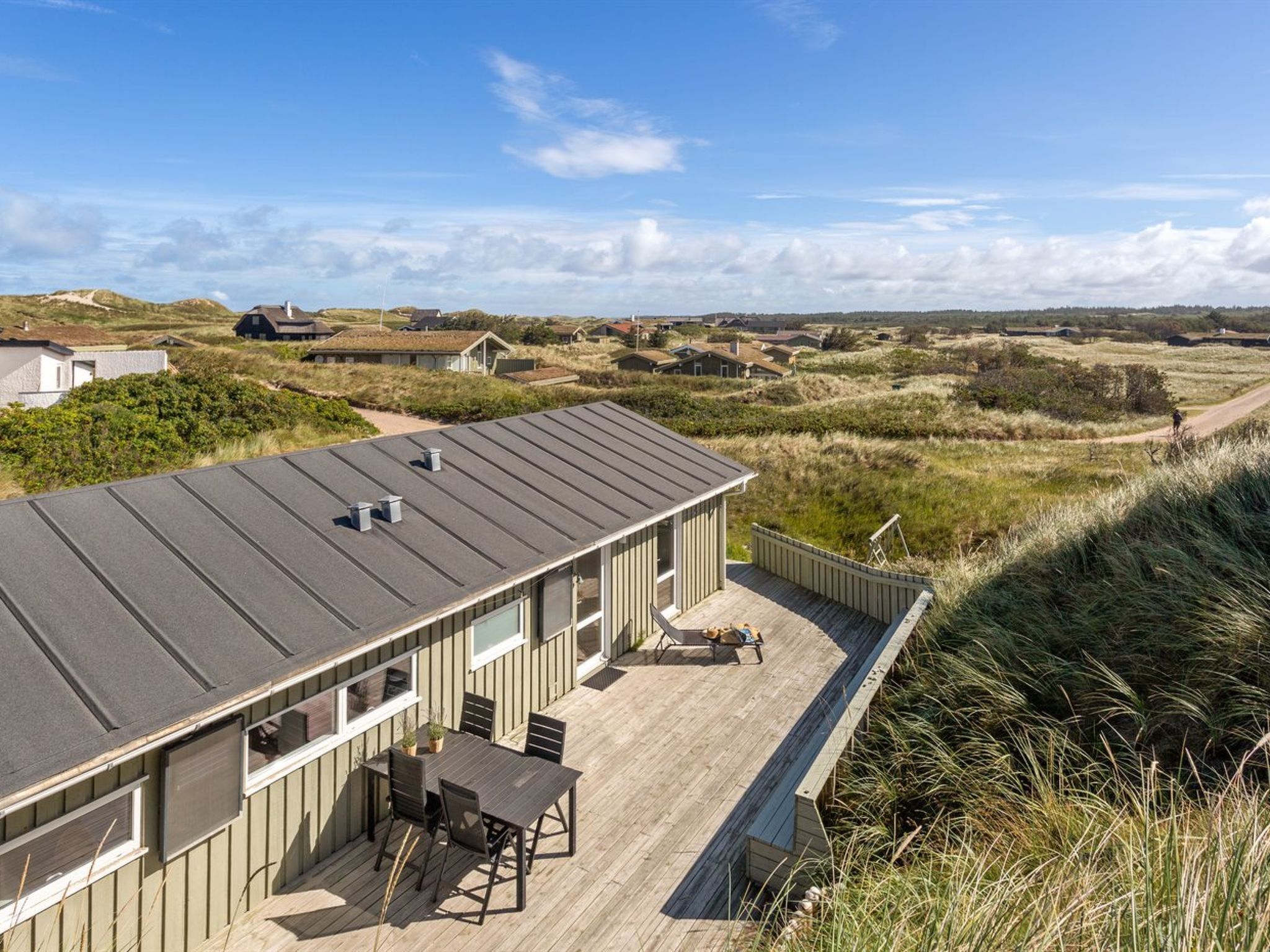 A modern house with a flat roof, a deck with a table and chairs, and a view of the surrounding landscape.
