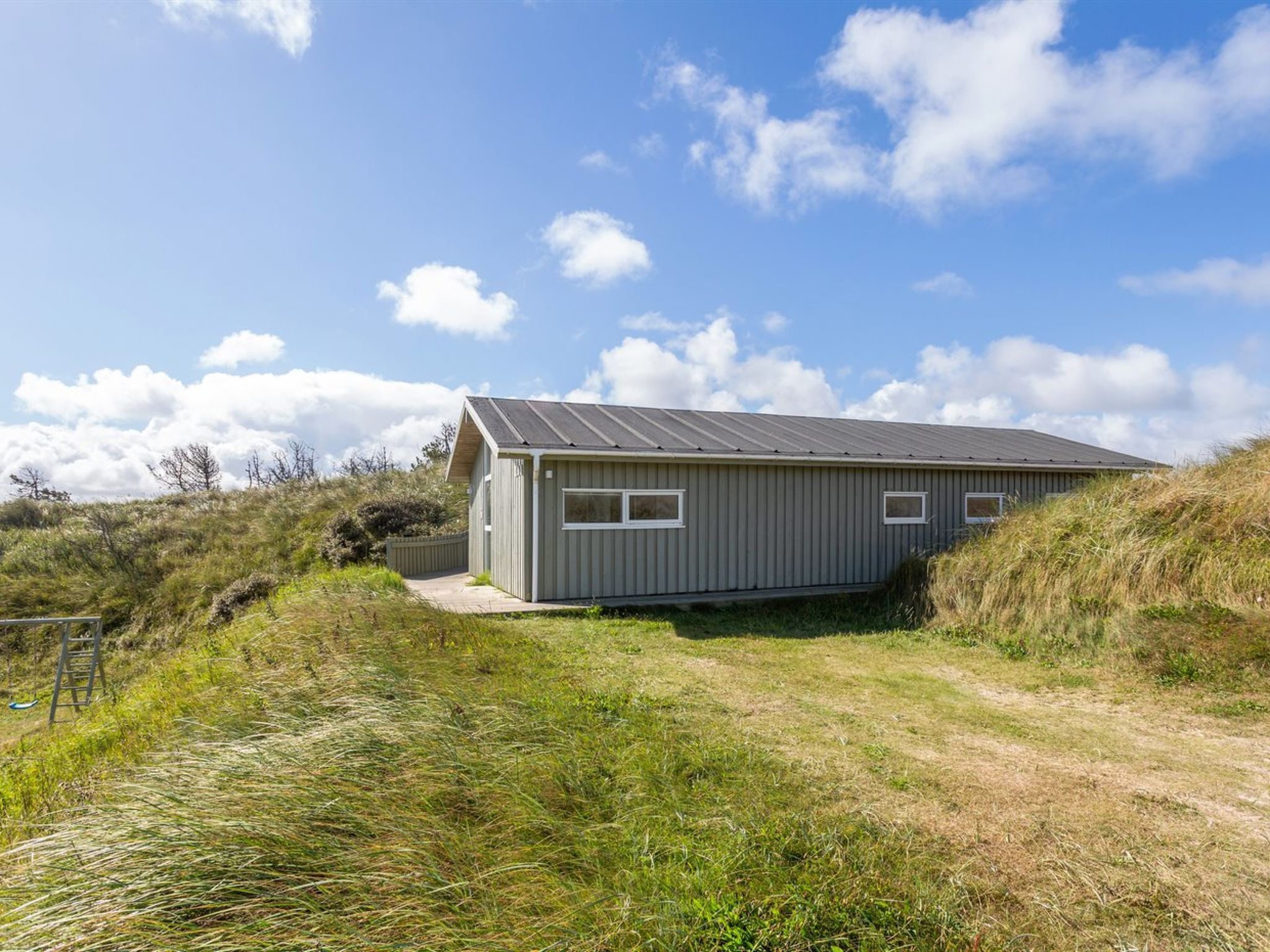 A single-story building with a sloped roof, situated on a grassy hill with a dirt path leading to it.