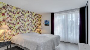Una habitación de hotel con cama, dos mesitas de noche, una silla, ventana con cortinas traslúcidas y papel tapiz con motivos florales.