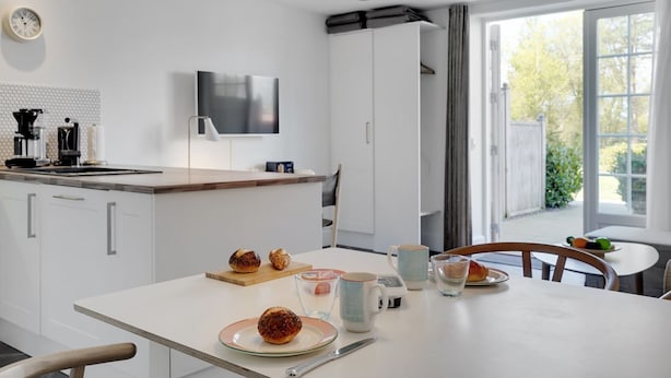 Una cocina moderna con armarios blancos, una mesa de comedor con alimentos para el desayuno y un ventanal con vistas a la vegetación.