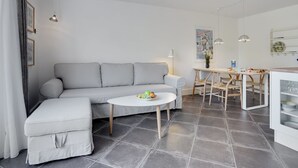 Una sala moderna con un sofá gris, una pequeña mesa redonda con frutas, una zona de comedor con una mesa blanca y sillas de madera, y una cocina con un armario blanco y estanterías.