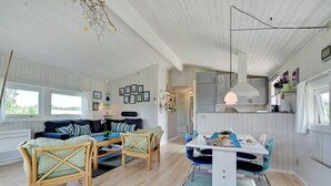 Een ruime woonkamer met een bank, een eettafel en stoelen. Er is een keuken met kasten en een hanglamp.