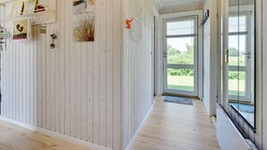 Een ruime kamer met houten vloeren en wanden, een grote glazen deur die uitkomt op een tuin, en diverse decoraties aan de muren, waaronder ingelijste foto's en een bord met een haan erop.