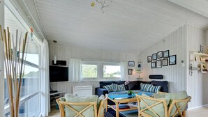 Een ruime woonkamer met een grote zithoek, twee rieten fauteuils en een flatscreen televisie aan de muur.