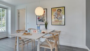 Zona pranzo con tavolo bianco, sedie di legno e lampadario. Si notano due poster incorniciati al muro e una finestra con vista su piante.