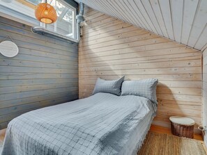 A bedroom with a sloped ceiling, wooden walls, a bed with a checkered blanket, a round wall clock, and a wicker stool.
