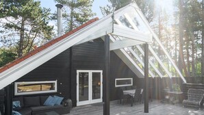 A modern black cabin with a sloped roof, a deck with outdoor furniture, and a large window overlooking the surrounding forest.