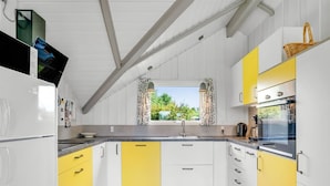 Una cocina con armarios blancos y amarillos, piso de madera y ventana con cortinas.