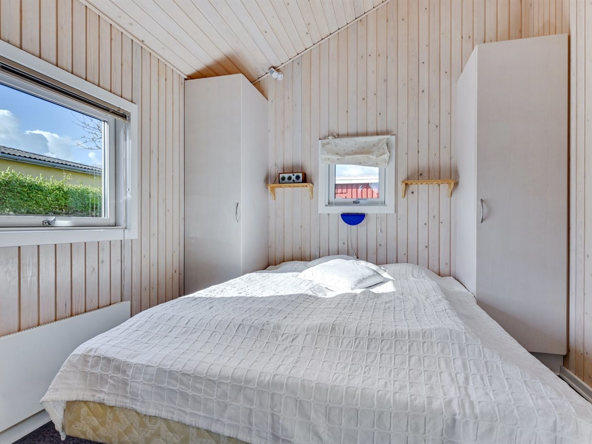 A bedroom with a bed, a window, a wardrobe, and a small shelf with a radio.