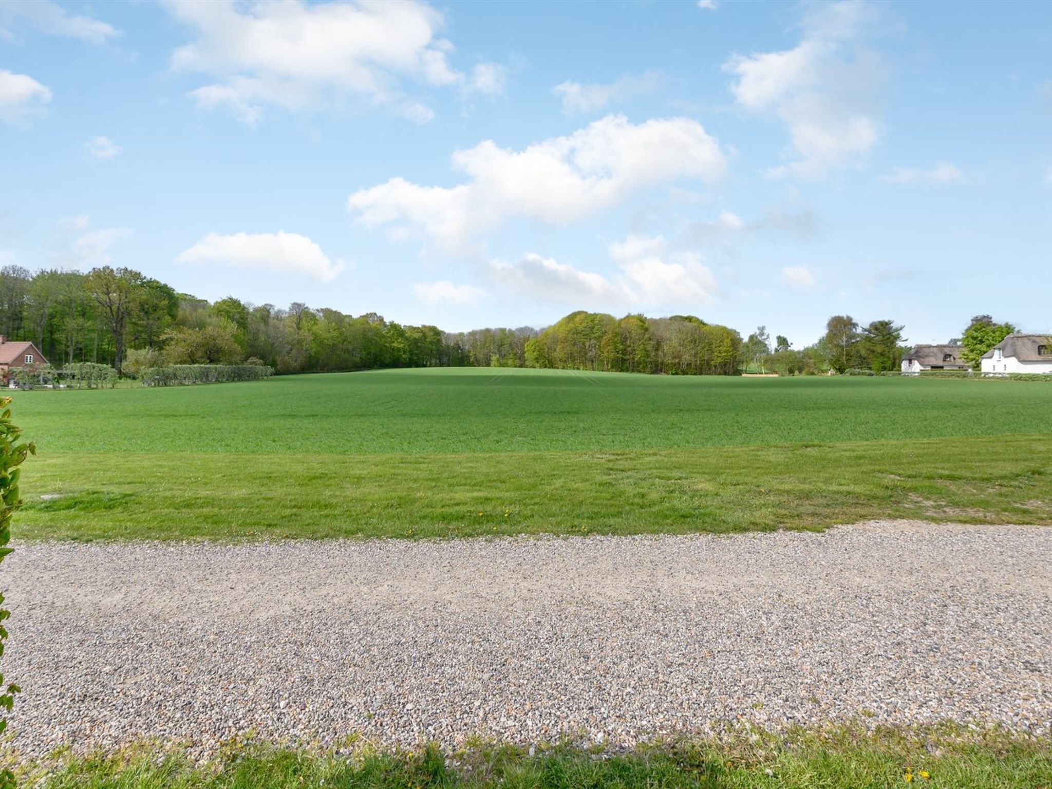 Un vaste champ ouvert, avec un chemin de gravier au premier plan et un groupe de maisons à l’arrière-plan.