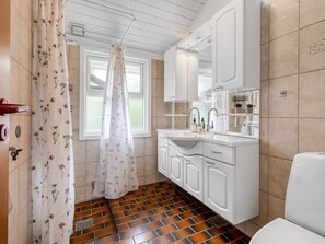 Une salle de bain avec un lavabo blanc, un miroir et des armoires, un rideau de douche à motifs floraux et une fenêtre donnant sur un paysage agréable.