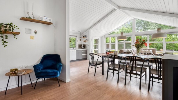 Een ruime keuken met witte kasten, een eethoek met houten stoelen, en een groot raam met uitzicht op groen.