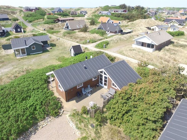 A coastal house with a sloped roof, a patio with outdoor furniture, and a view of the surrounding houses and dunes.