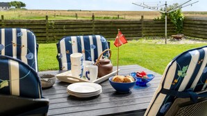 Une table à manger installée à l’extérieur, avec des chaises à rayures, un drapeau, une bouilloire, des tasses, des assiettes et un saladier rempli de fruits.