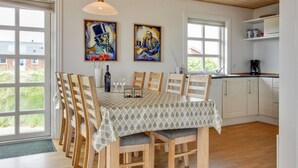 En spiseplads med et bord dækket til seks personer, træstole og en borddug med mønster. Væggene er prydet med to malerier, et vindue med udsigt til grøn bevoksning, og et køkkenområde med skabe og en kaffemaskine.