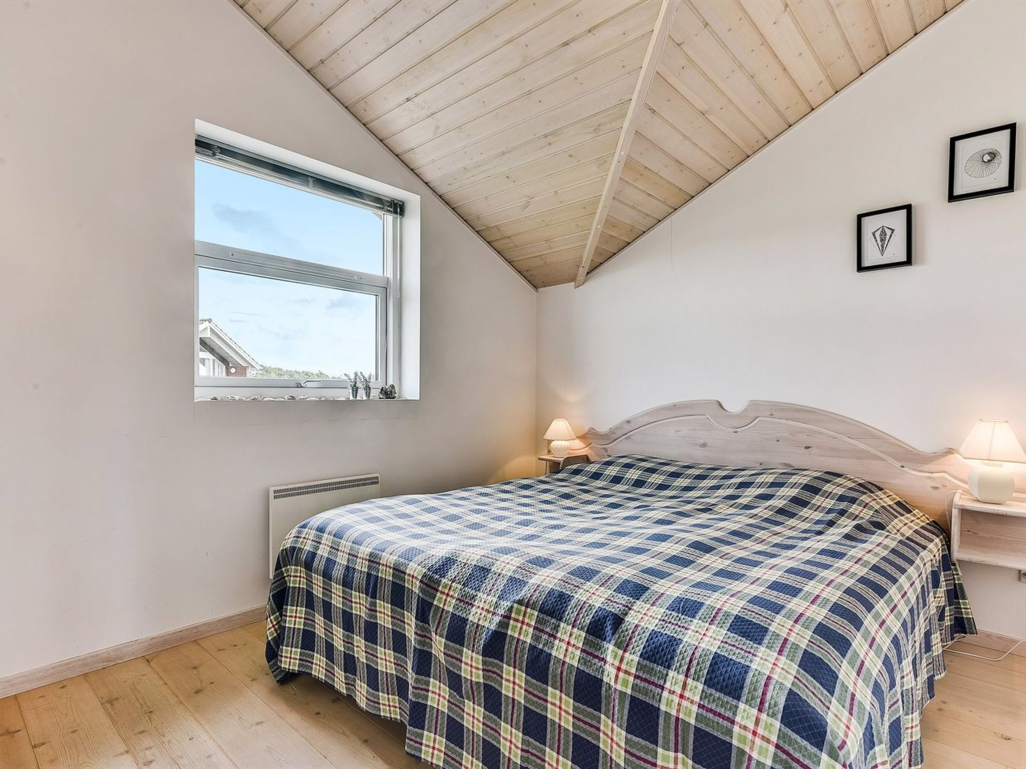 A bedroom with a sloped ceiling, a window, a bed with a plaid bedspread, two bedside lamps, and a radiator.