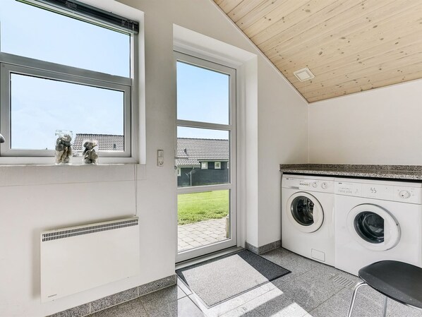 A laundry room with two washing machines, a radiator, and a window with a view of a house and lawn.