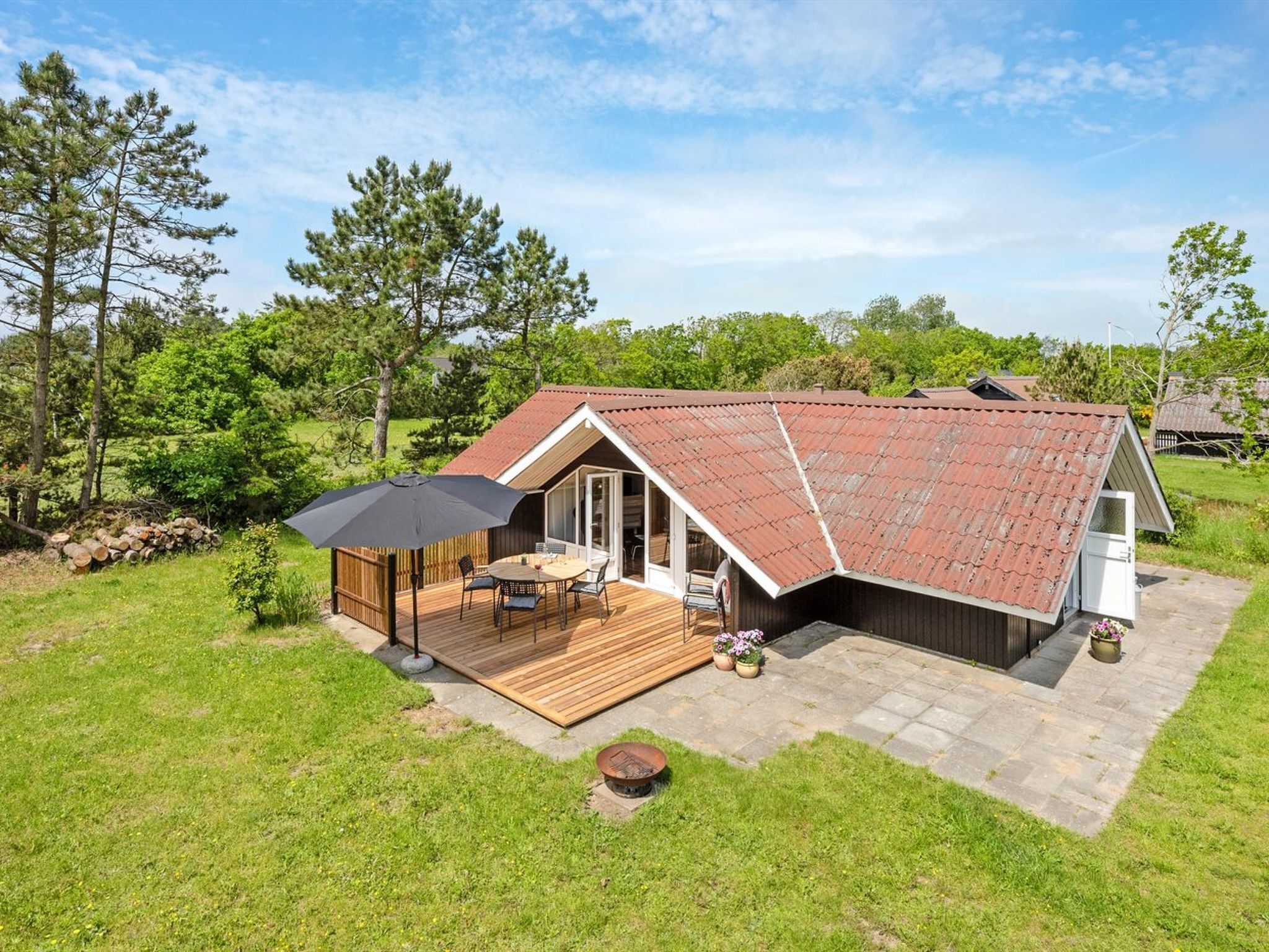 Eingeschossiges Haus mit geneigtem Dach, einer Terrasse mit Gartenmöbeln und einer Feuerstelle.