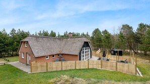 Une maison à toit de chaume, avec une cour clôturée, un petit abri de jardin et un banc.