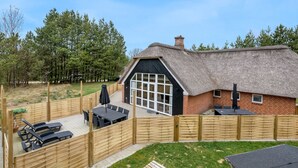 Un chalet au toit de chaume, doté d’une grande fenêtre, d’une terrasse avec des meubles d’extérieur et d’une clôture en bois.