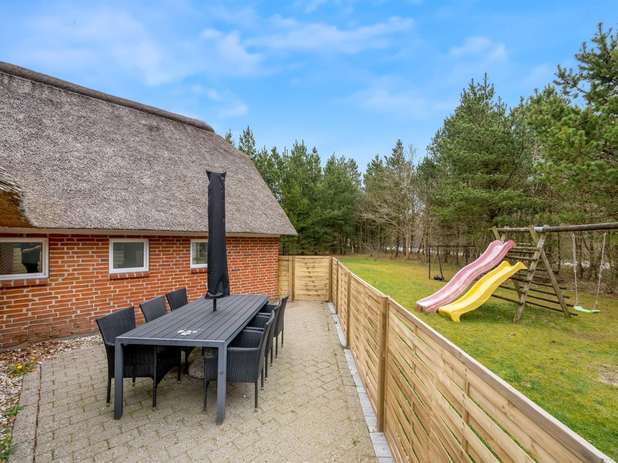 A brick building with a thatched roof, a patio table with chairs, and a wooden fence enclosing a grassy area with a playground.