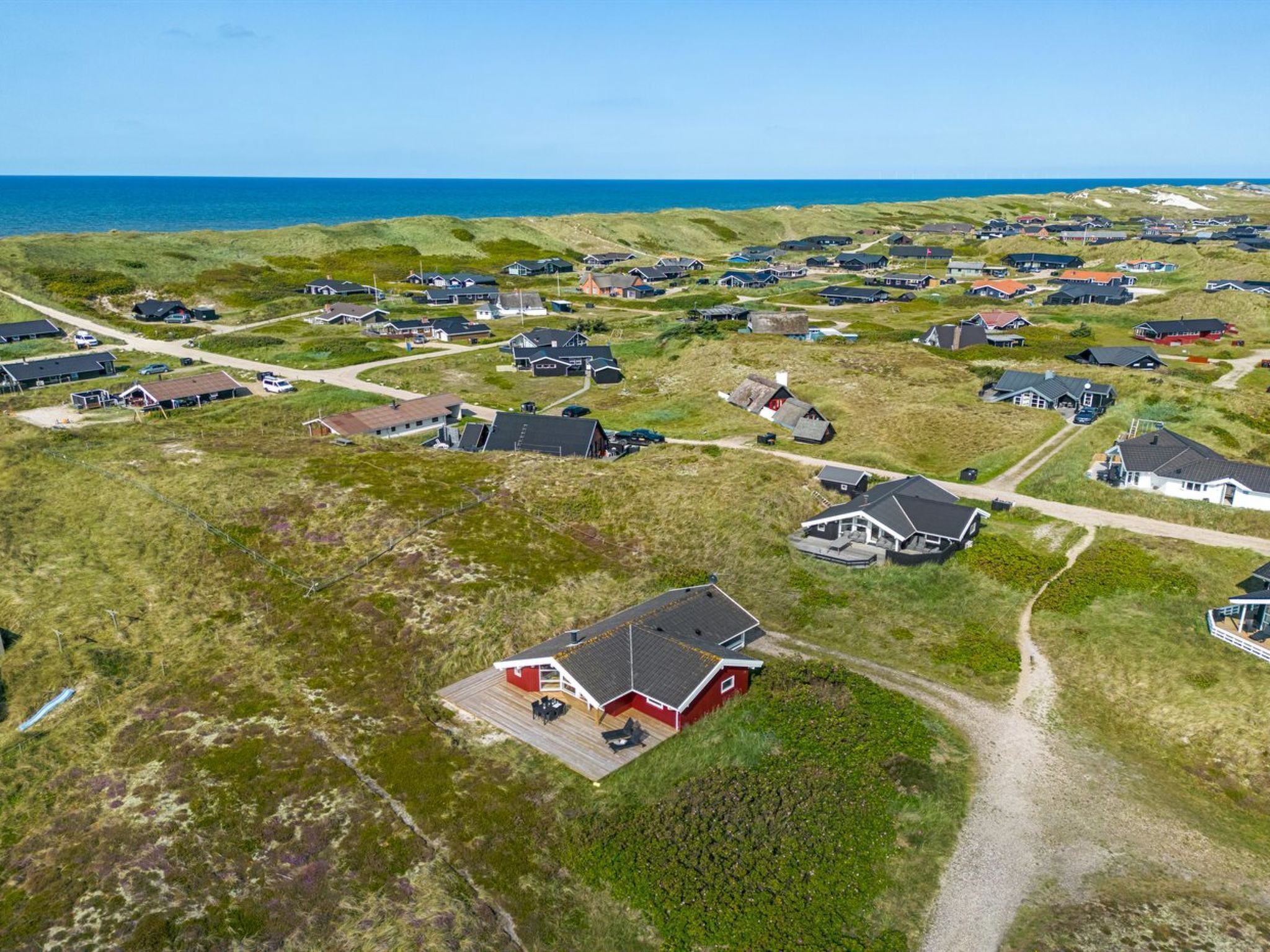 An aerial view of a coastal village with houses spread across grassy dunes, connected by narrow roads, and overlooking the sea.