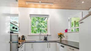A modern kitchen with white cabinets, a window with a view of greenery, and a wooden ceiling.